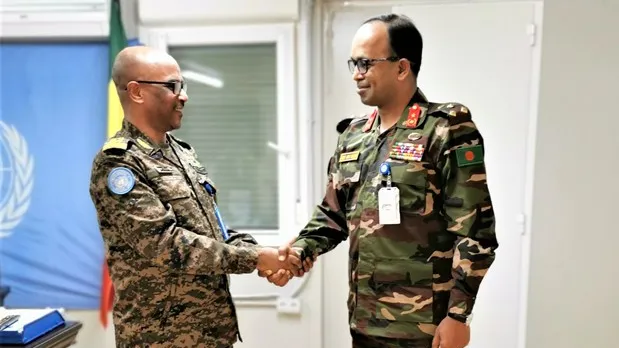 Two men in military uniforms standing in a room shaking hands. In the background, there is a UN flag.
