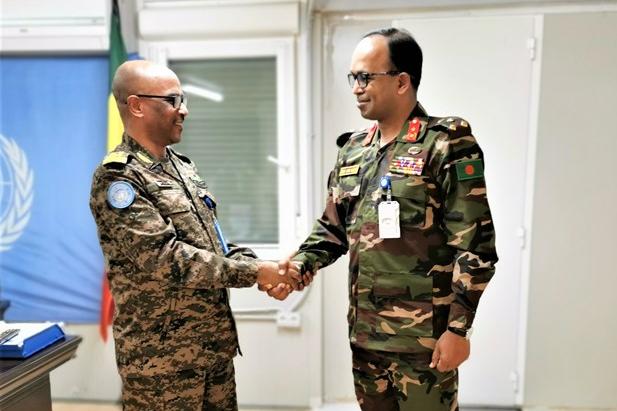 Two men in military uniforms standing in a room shaking hands. In the background, there is a UN flag.